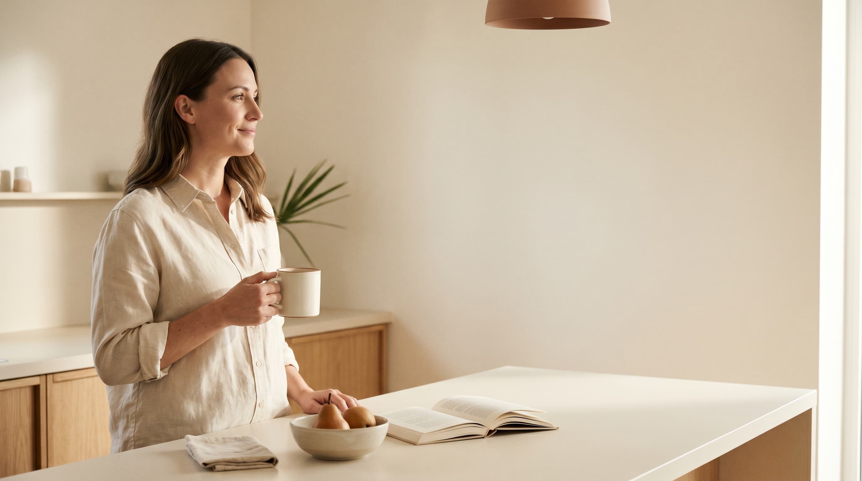 Woman in her late thirties at a warm cream kitchen island in mid-morning light, mug in hand, shoulders relaxed